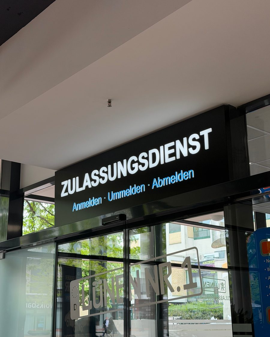 Beleuchtete Werbeanlage „ZULASSUNGSDIENST" von außen mit Blick auf den Eingang und die „LÜNEN NR.1"-Beschriftung an der Glasfassade