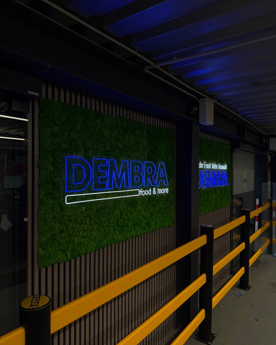 Diagonale Nahaufnahme des LED-Neon-Logos "DEMBRA food & more" auf der Mooswand im Eingangsbereich von DEMBRA Food & More