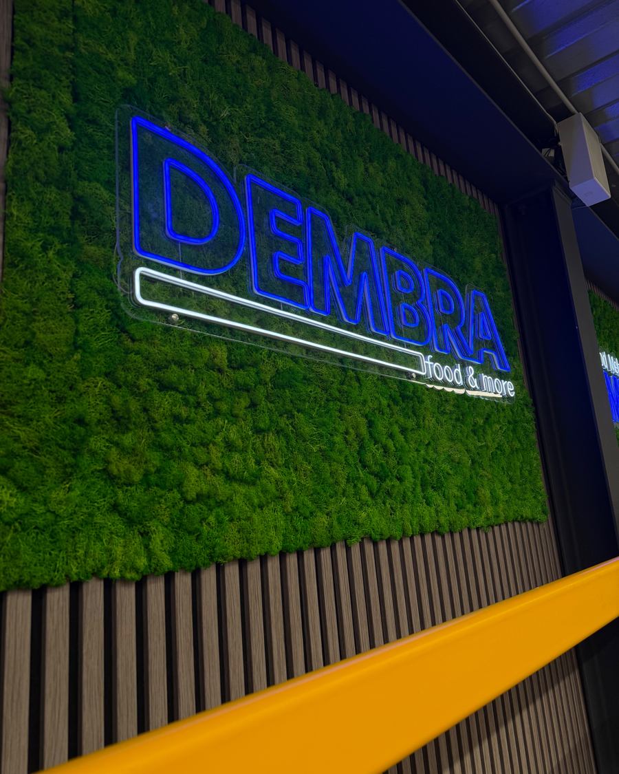 Diagonale Nahaufnahme des LED-Neon-Schriftzugs "Mehr Food Mehr Auswahl DEMBRA" auf echtem Moos
