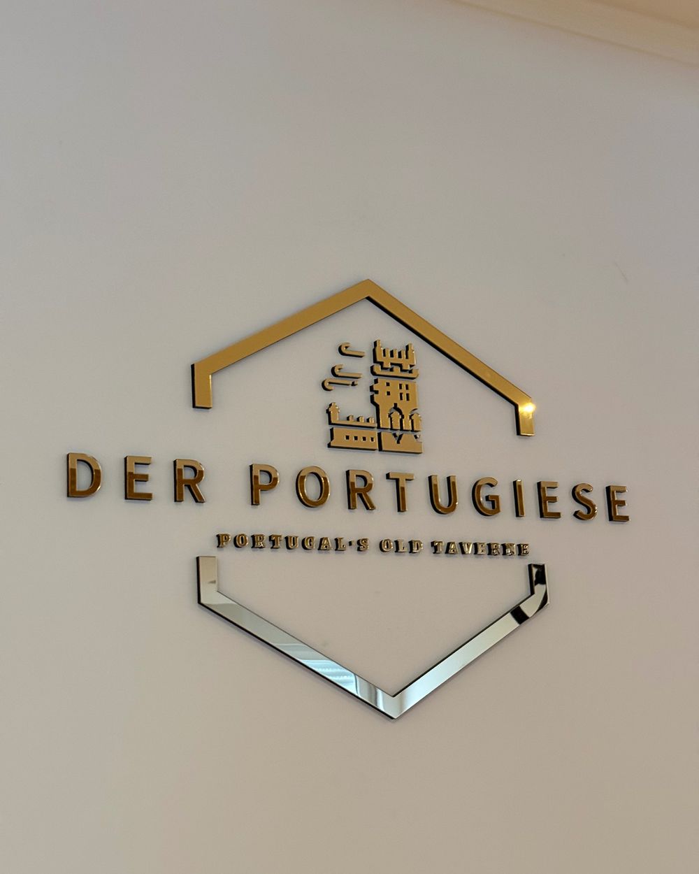 Goldenes 3D-Wandlogo "DER PORTUGIESE / Portugal's Old Taverne" mit Wappen- und Turm-Icon an einer weißen Innenwand