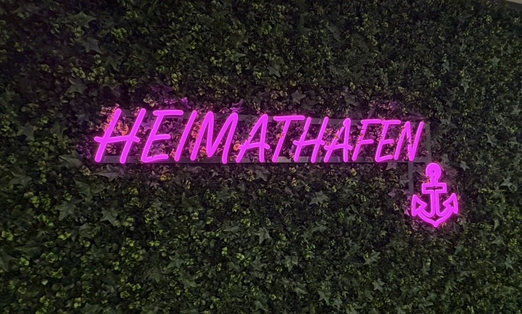 LED-Neon-Schild mit Schriftzug HEIMATHAFEN und Anker in Pink auf der Kunstpflanzenwand im Flur der Ärzteversorgung Westfalen-Lippe in Münster am Abend, gefertigt von Visuellewand