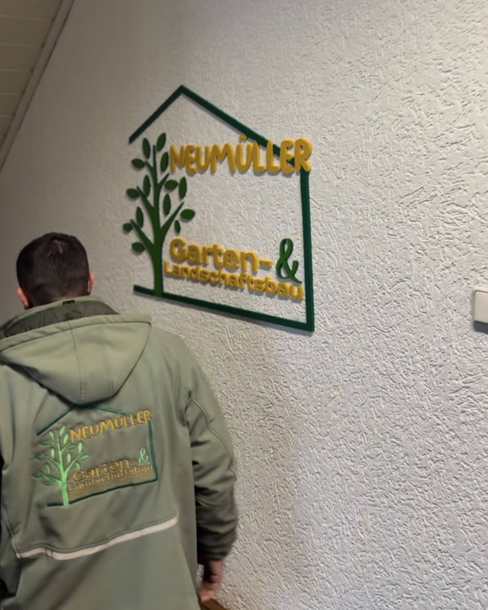 Mitarbeiter von GALA Bau Neumüller mit dem Firmenlogo auf der Arbeitsjacke geht auf das 3D-Wandlogo im Büro zu