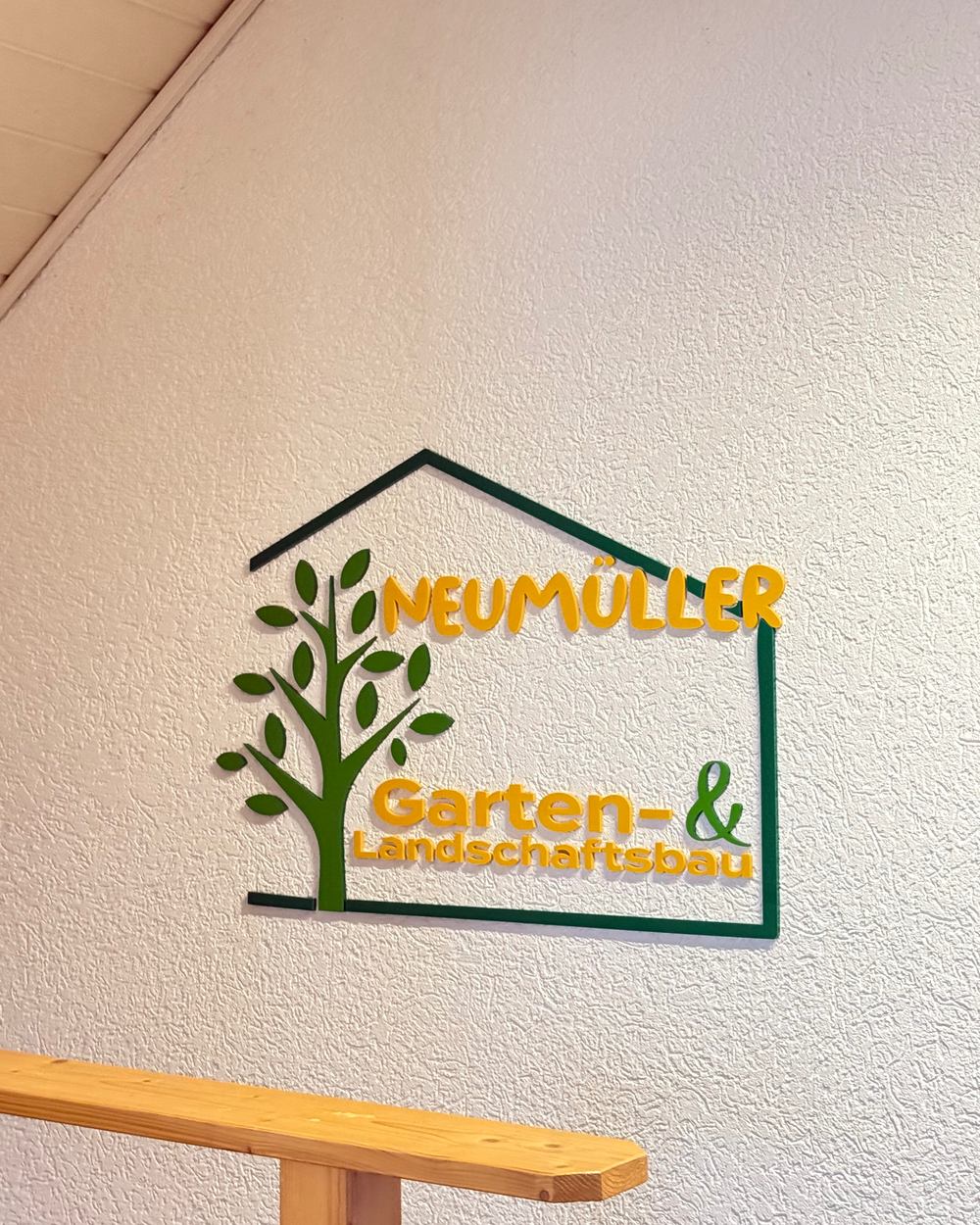 Nahaufnahme des 3D-Wandlogos von GALA Bau Neumüller (Garten- und Landschaftsbau) mit Hausform, Baum-Symbol und Schriftzug