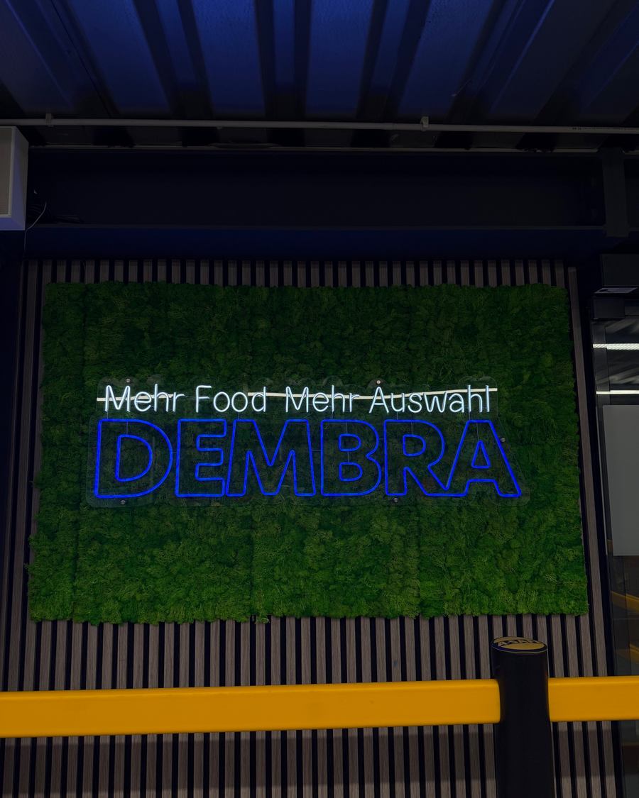 Nahaufnahme des blau leuchtenden LED-Neon-Schriftzugs "DEMBRA food & more" auf echtem Moos, Dortmund