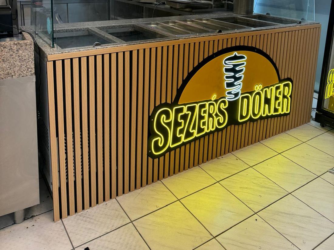 Seitliche Ansicht des beleuchteten „Sezer's Döner"-Logos auf der Holzlamellen-Thekenfront, im Hintergrund die Speisekarten-Bildschirme und der Dönerspieß-Grill